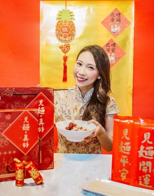 一名女子拿著一碗老薑麻油麵線，旁邊是錯落的大紅色禮盒及提袋。