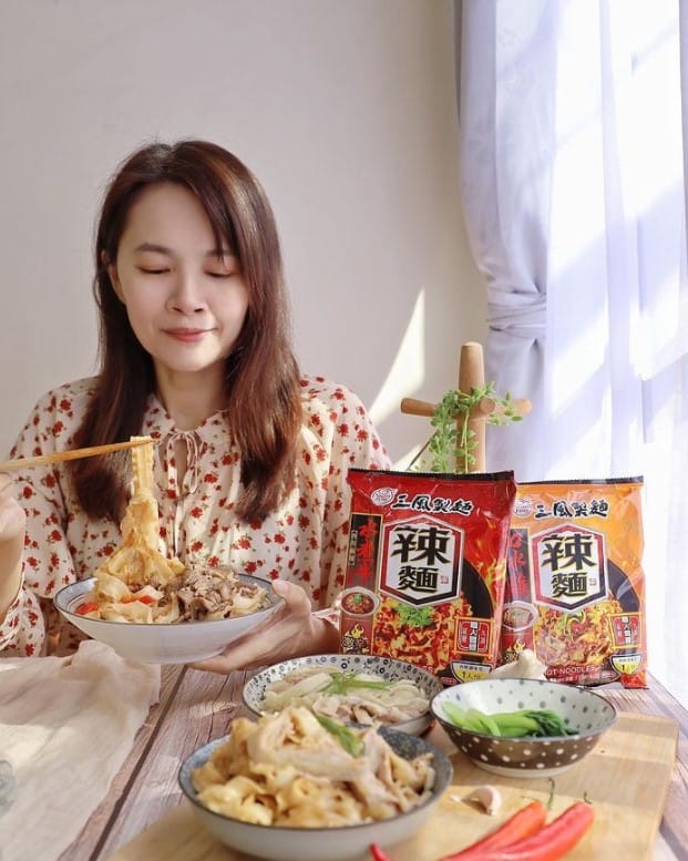 一名女性手裡捧著一碗麵，旁邊擺放著不同配菜與這碗乾拌麵的外包裝。